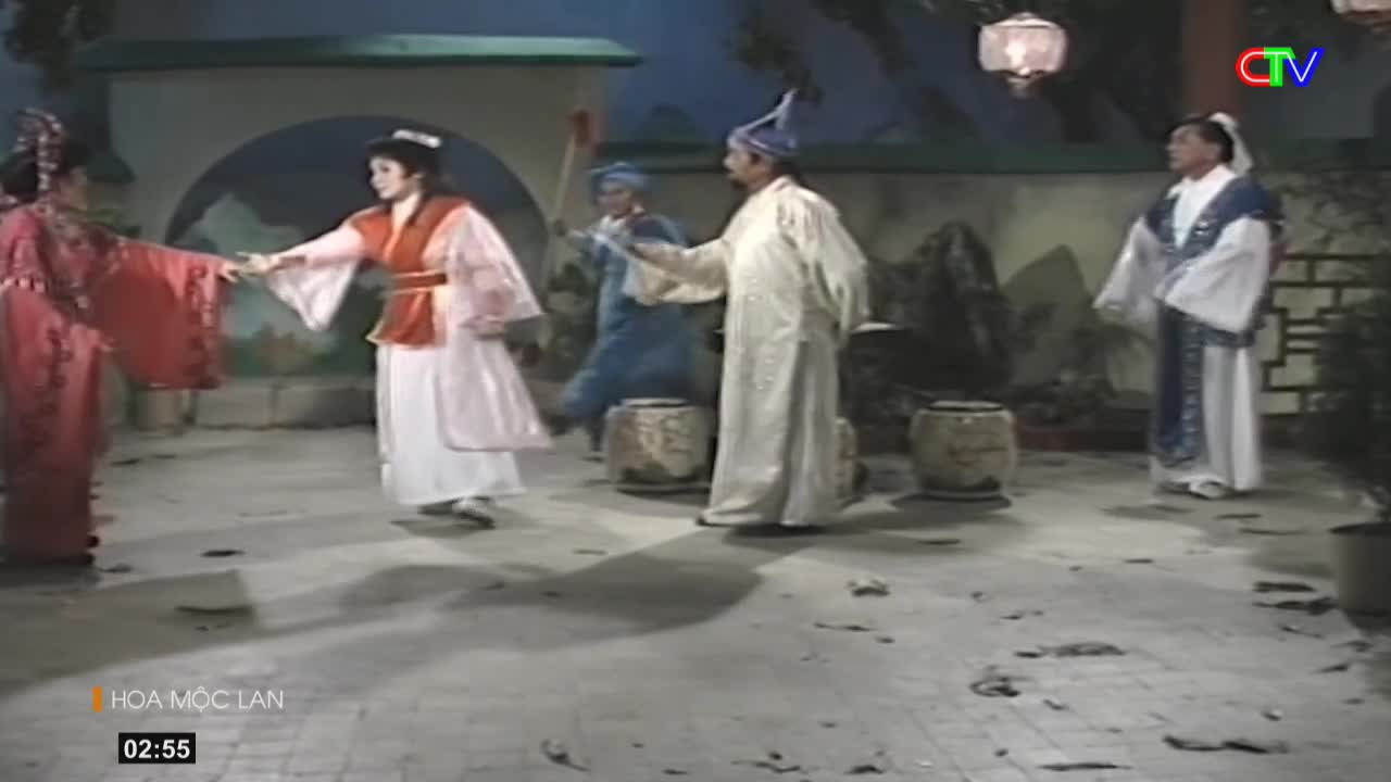 Two women in flowing robes, one in vibrant red and the other in white and orange, clasp hands. Behind them, men in traditional Vietnamese attire stand near large ceramic pots.