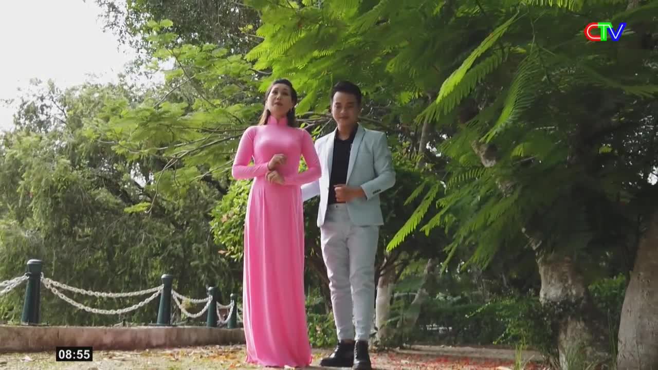 A woman in a bright pink áo dài stands beside a man in a light blue suit. They are positioned in front of lush green trees, with a white chain fence behind them.