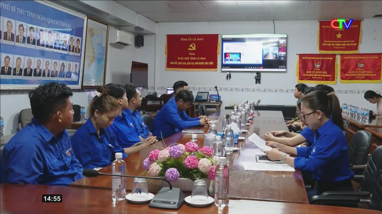 A group of young people in blue shirts are gathered around a long table, some looking at papers, others at a screen. A banner for Ca Mau TV hangs on the wall behind them.