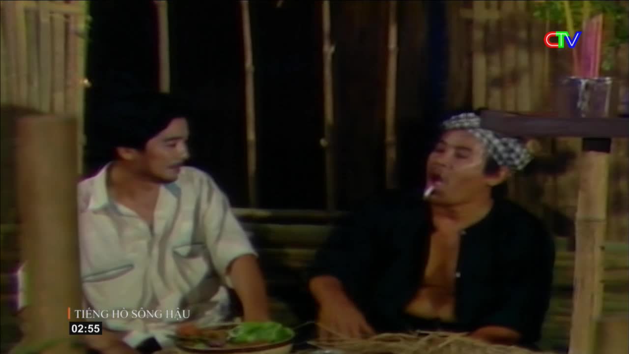 A man in a white shirt leans forward, his gaze fixed on something to his right. Beside him, another man, wearing a checkered headscarf, exhales smoke from a cigarette held between his lips.