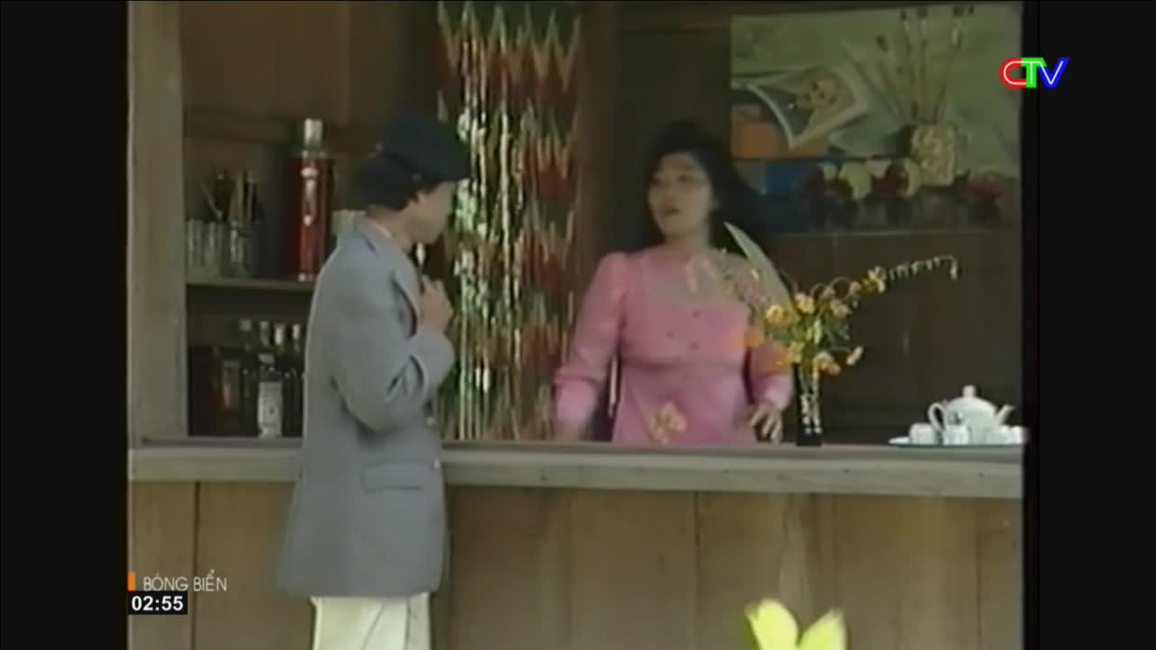 A man in a light grey jacket stands at a wooden counter, facing a woman in a pink dress. The woman gestures with her hands as she speaks, her dark hair falling around her shoulders. Behind them, shelves hold bottles and glasses, and a vase of yellow flowers sits on the counter.