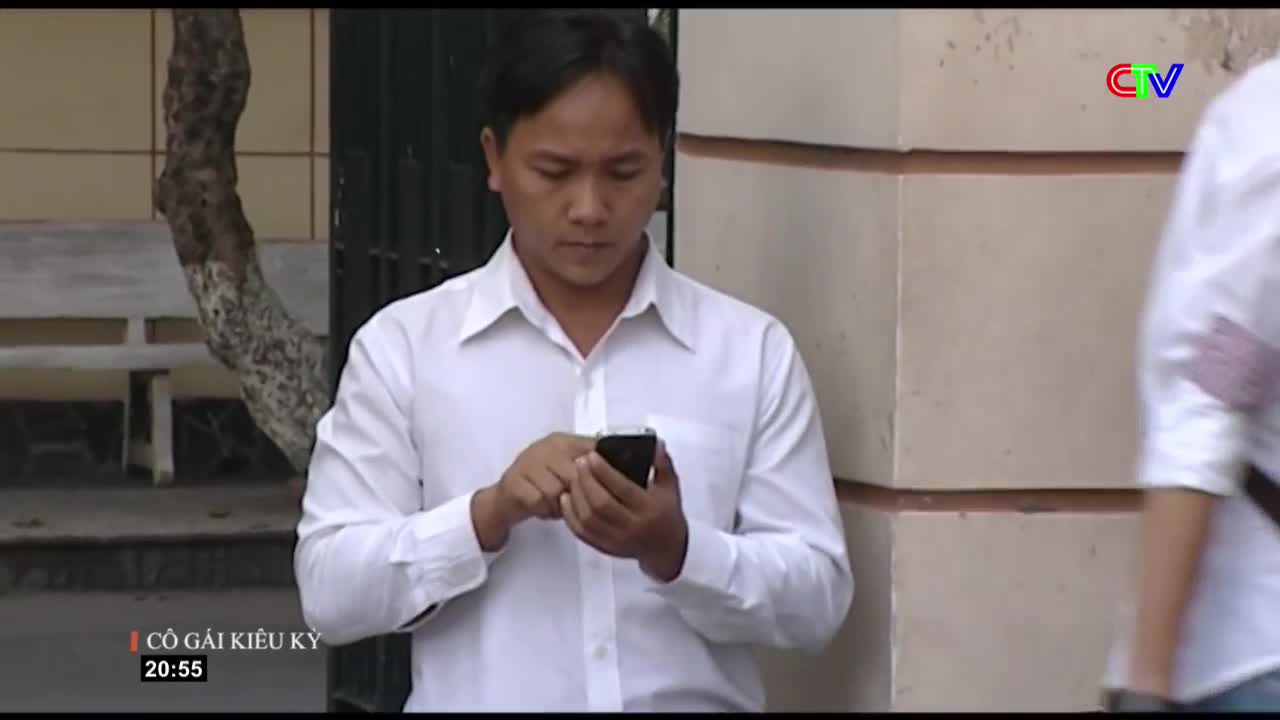 A man in a white shirt is focused on his phone, his thumbs moving across the screen. Another person in a white shirt walks past in the background, their form slightly blurred.