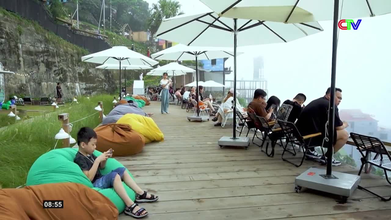 A young boy sits on a bright green beanbag, absorbed in his phone. People lounge on beanbags and chairs under large white umbrellas on a wooden deck, overlooking a misty landscape that hints at Vietnam.