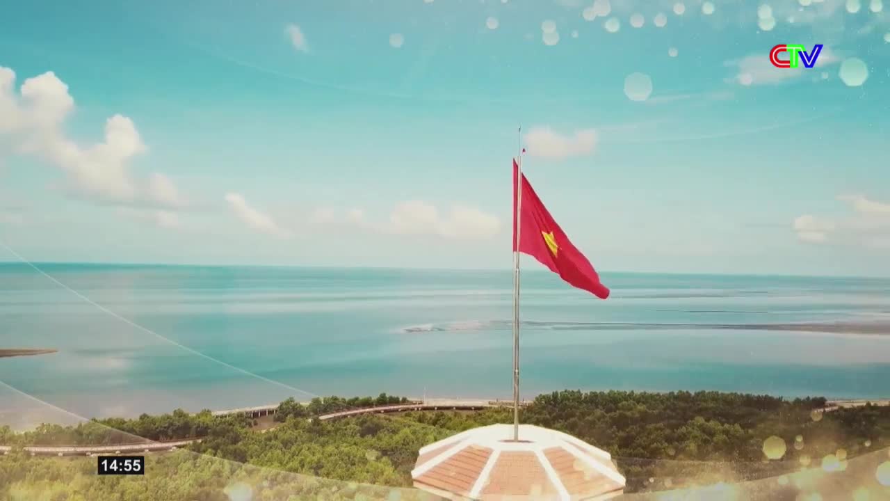 The Vietnamese flag snaps in the breeze against a vast, calm sea. A bridge snakes through green trees in the distance.