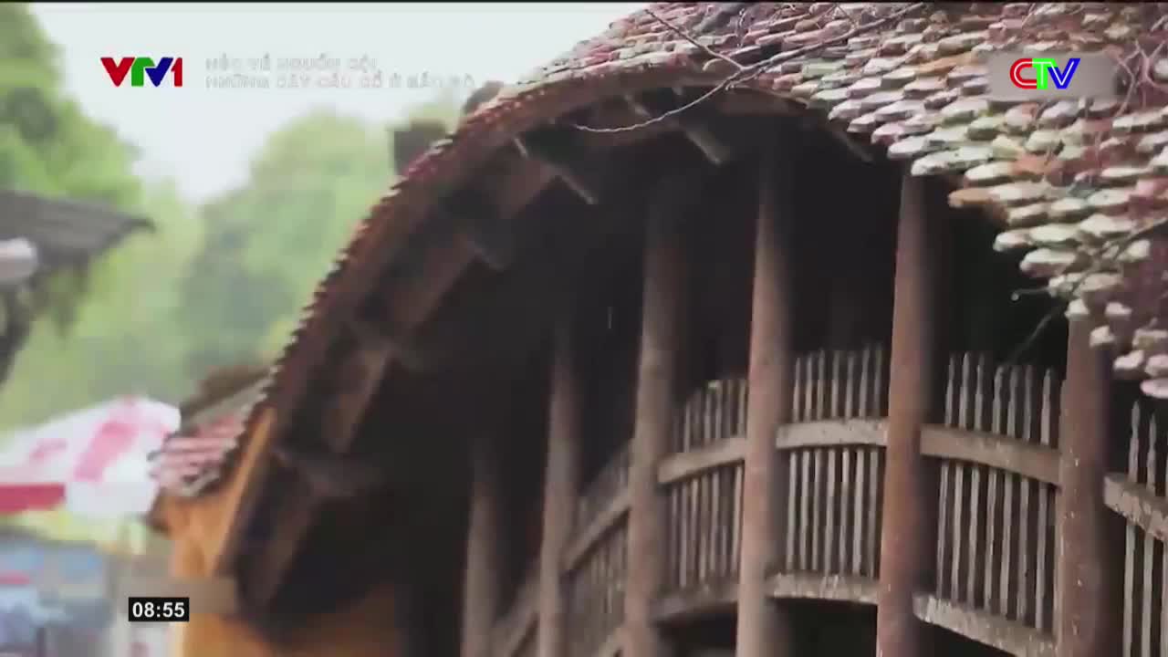 Raindrops fall on the weathered tile roof of a traditional wooden structure. A red and white umbrella is visible in the background, suggesting a market or street scene in Vietnam.