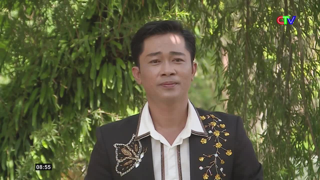 A man in a black jacket with embroidered gold flowers and a butterfly stands before lush green foliage. The Ca Mau TV logo appears in the upper right corner.