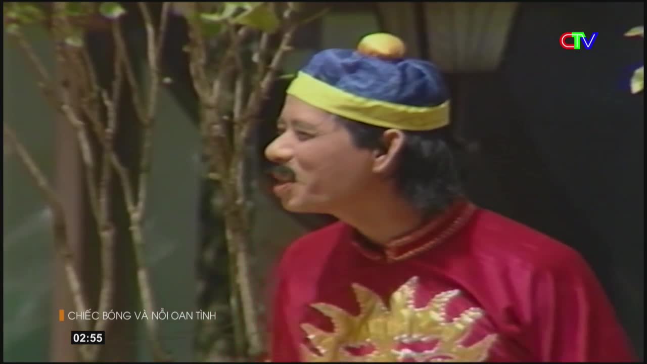 A man in a bright red tunic and a blue and yellow hat with a yellow pom-pom speaks animatedly. The Ca Mau TV logo appears in the upper right corner.