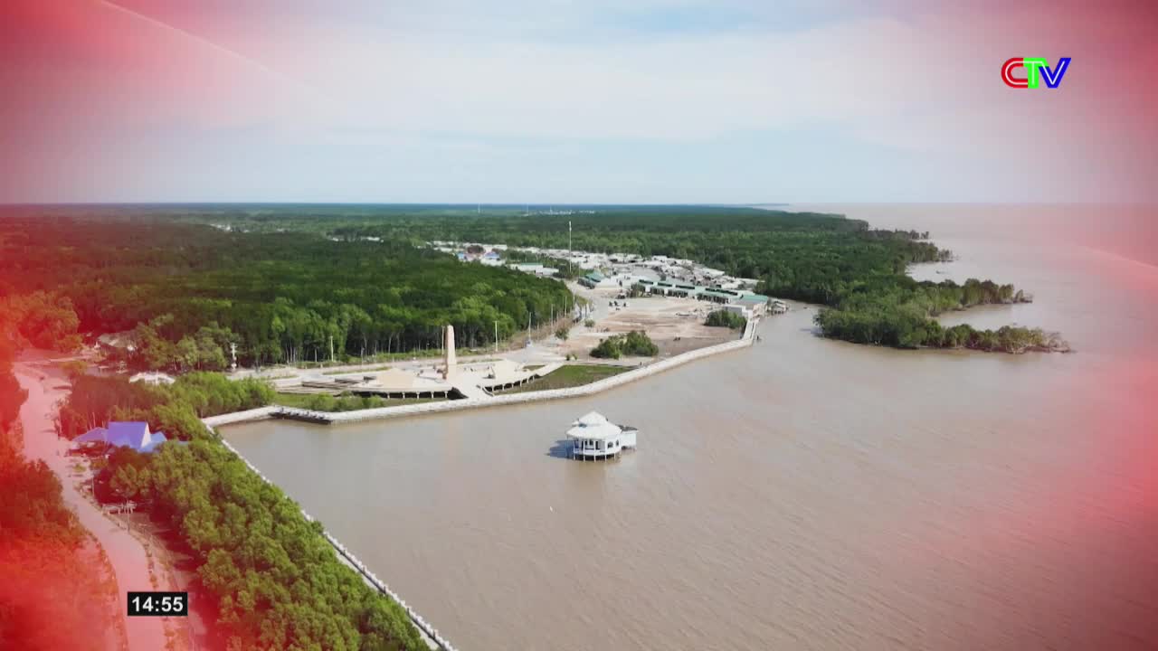 The brown water of the Ca Mau river flows past a white, stilted building. Mangrove trees line the shore, leading to a cluster of buildings and a tall monument.