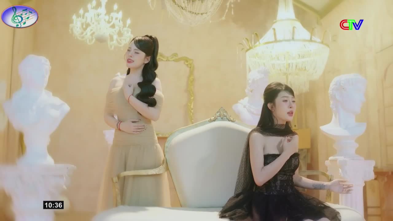 Two young women sing in a gilded room, bathed in soft light. One stands in a flowing tan dress, the other sits in a dark, ornate gown, both with hands pressed to their chests.