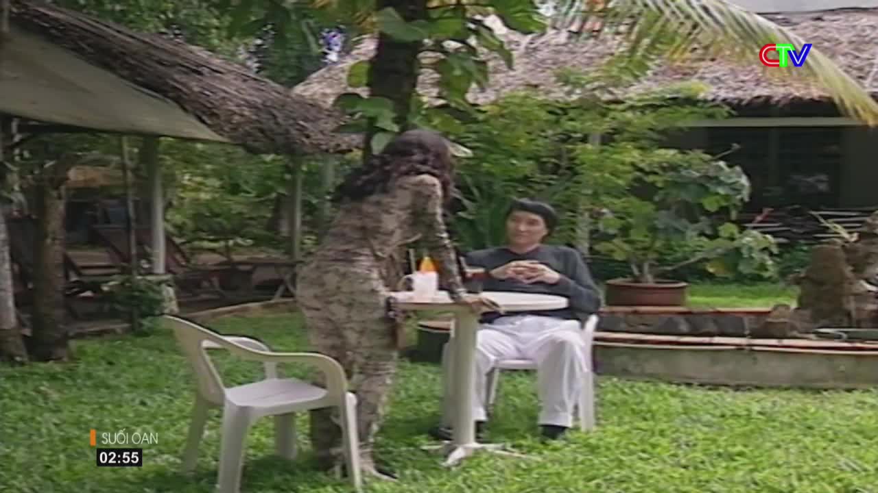 A woman in a patterned dress leans over a table, talking to a man seated in white pants. The scene unfolds in a lush, green garden, likely in Ca Mau, with a thatched roof structure visible.