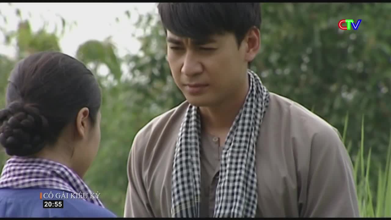 A man in a tan shirt and checkered scarf looks down with a troubled expression. Behind him, tall green reeds sway gently in the breeze.