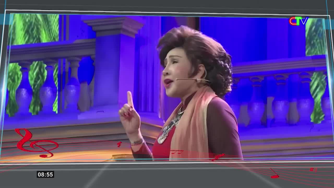 A woman with a dramatic hairstyle and a peach-colored scarf sings into a microphone, her right index finger raised. Behind her, a stage set features blue lighting and ornate railings, reminiscent of a balcony. The Ca Mau TV logo appears in the upper right corner.