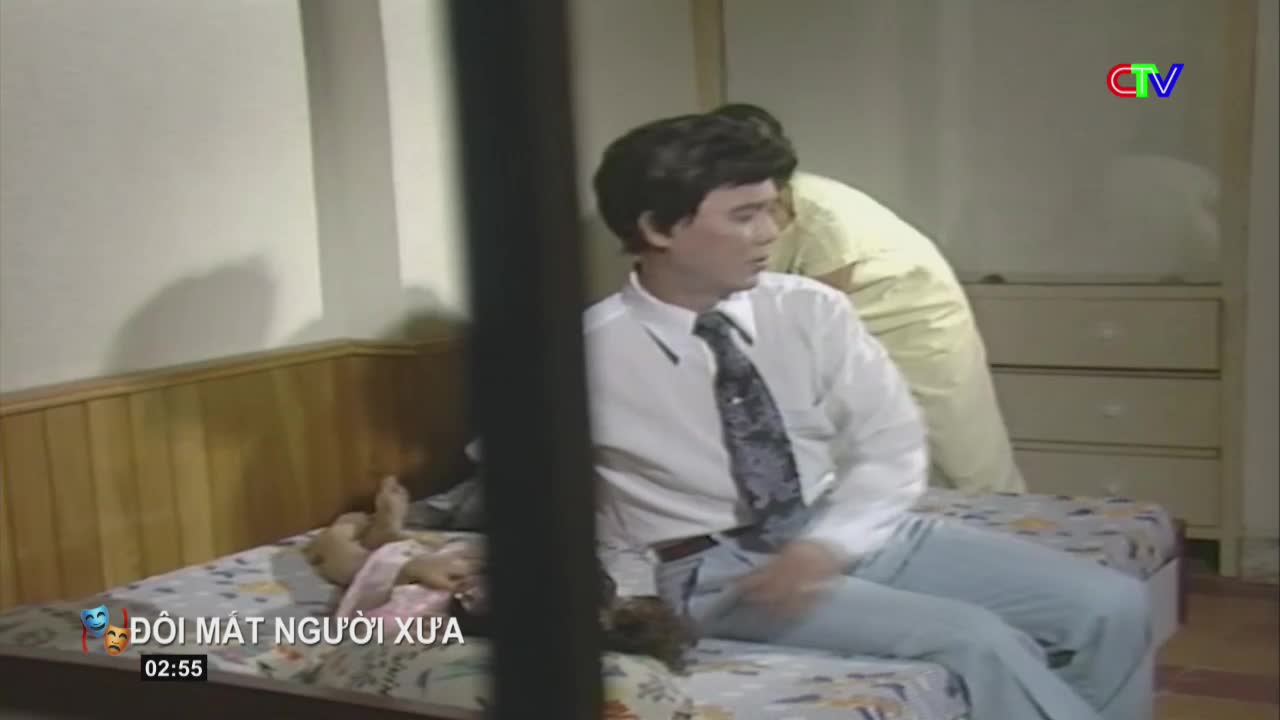 A man in a white shirt and tie sits on a bed, looking away. Another person, wearing yellow, stands behind him.