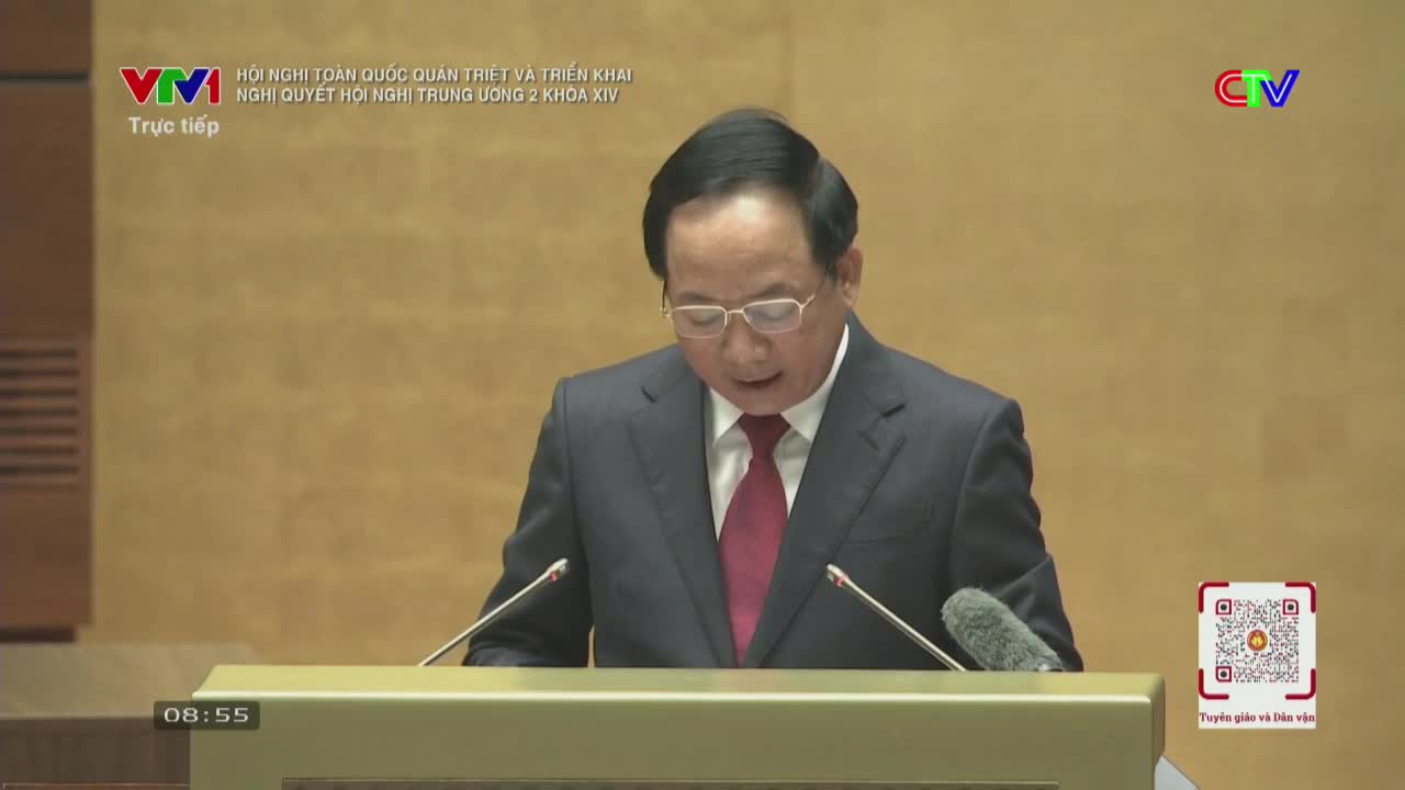 A man in a dark suit and red tie stands at a podium, speaking into microphones. The VTV logo appears in the corner, indicating this broadcast is from Vietnam.