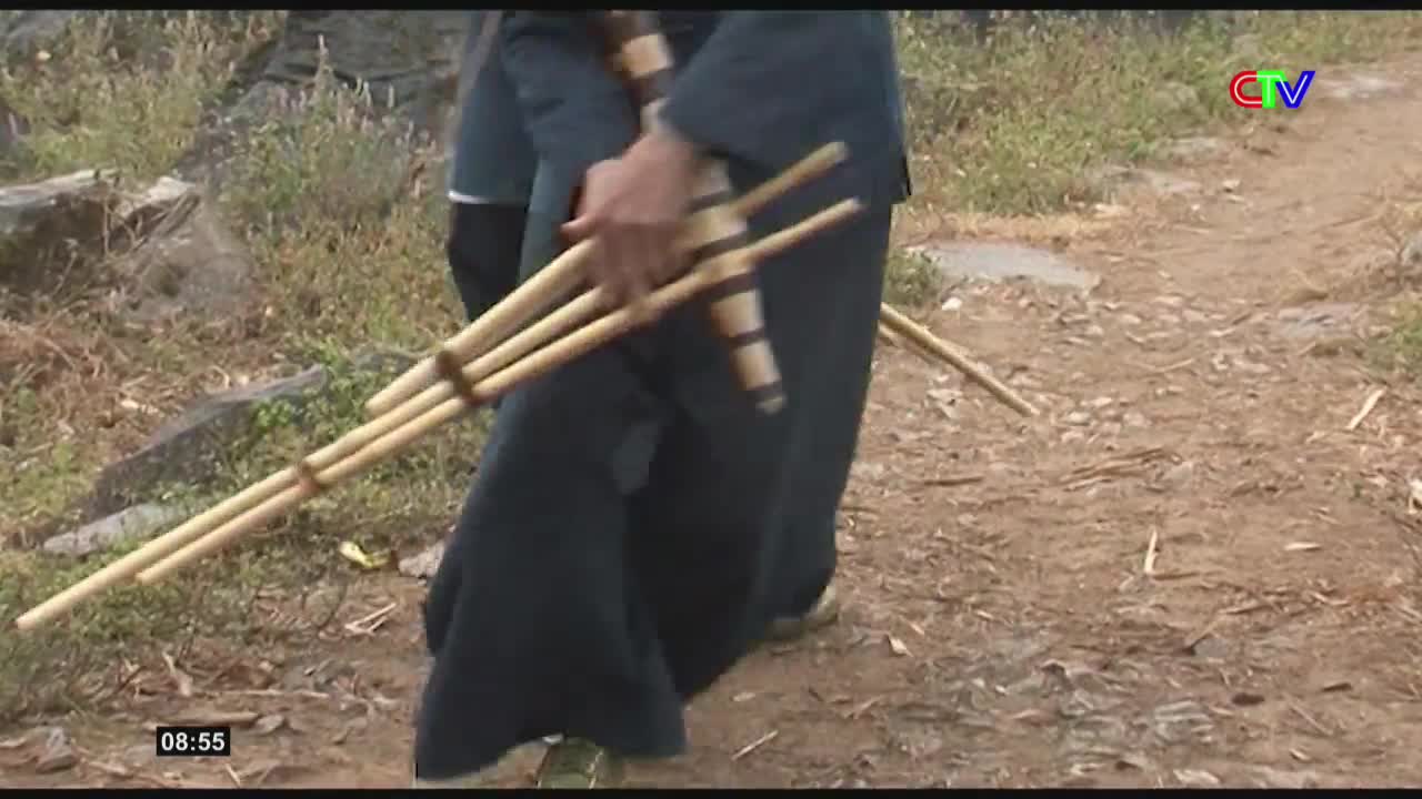 A person in dark clothing walks along a dirt path, carrying a bundle of bamboo pipes. The pipes are bound together with twine and extend out from their side.