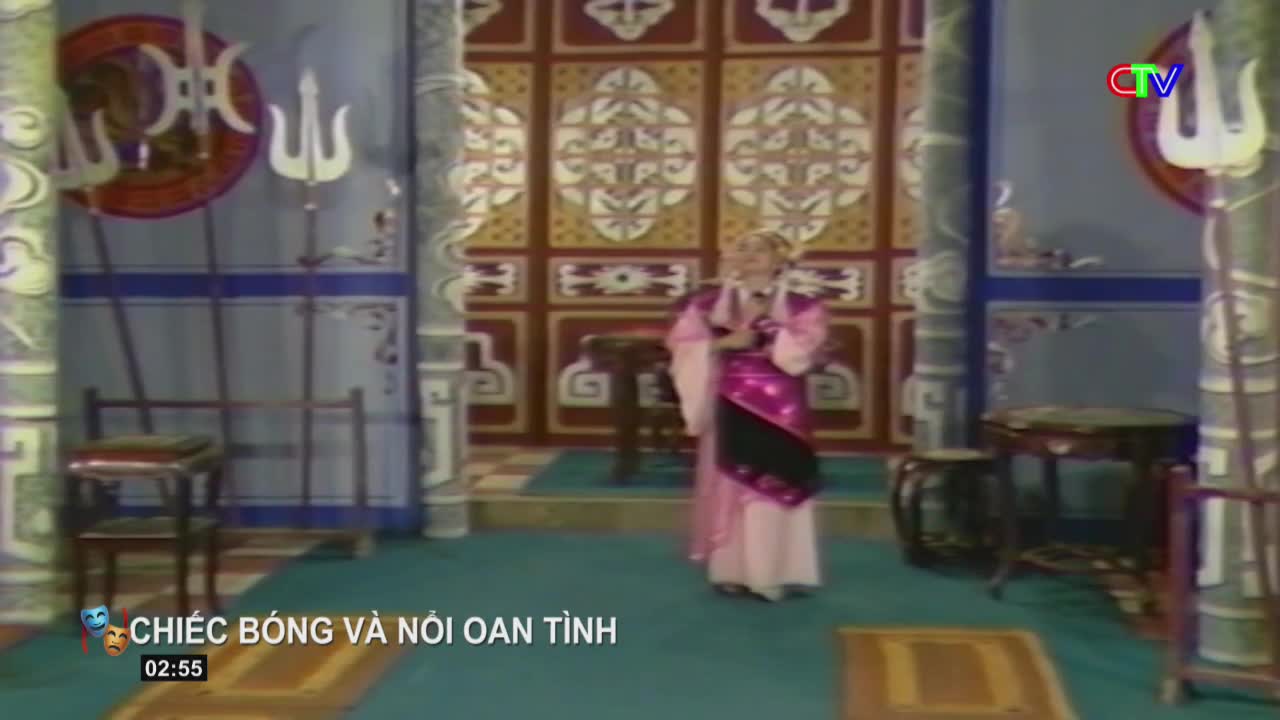 A performer in a pink robe stands on a stage, holding a prop. The backdrop is a richly decorated wall, typical of Vietnamese theater.