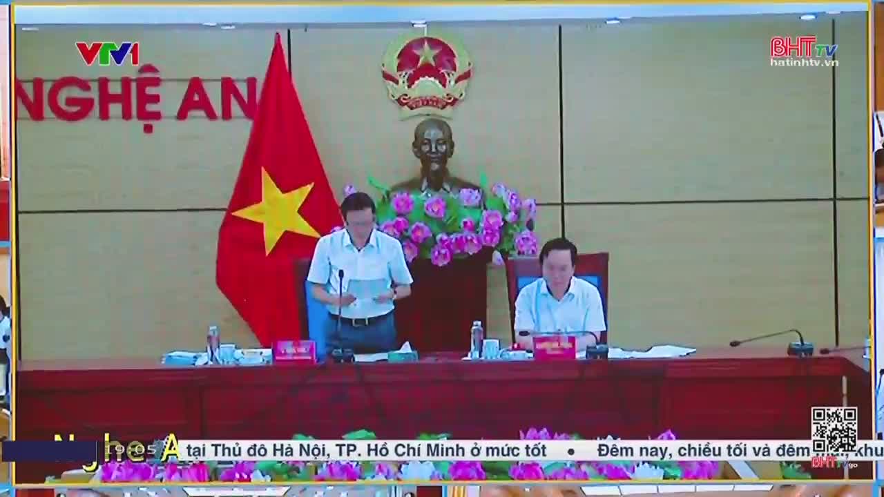 Two men in light-colored shirts sit behind a long table, reading from papers. A large Vietnamese flag and a bust of a man are visible behind them, as the Ha Tinh TV logo is displayed in the corner.
