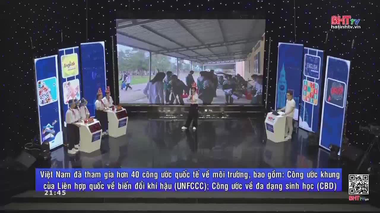 On the Ha Tinh TV stage, a presenter stands before a large screen showing a group of people under a covered area. Contestants in party hats sit at their podiums, ready to play.

