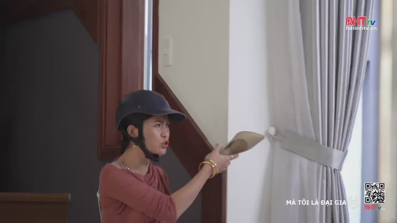 A woman in a helmet is holding a shoe, aiming it at the curtain. Her mouth is open, and she seems to be shouting, likely on a Ha Tinh TV show from Vietnam.
