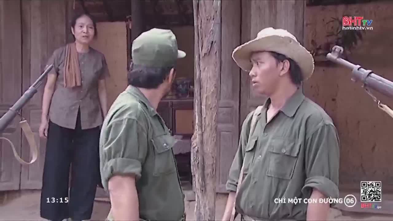 Two men in military-style shirts and hats are talking, one looking concerned, the other gesturing. A woman in a simple blouse and black pants stands nearby, a rifle resting against the wooden wall behind her.
