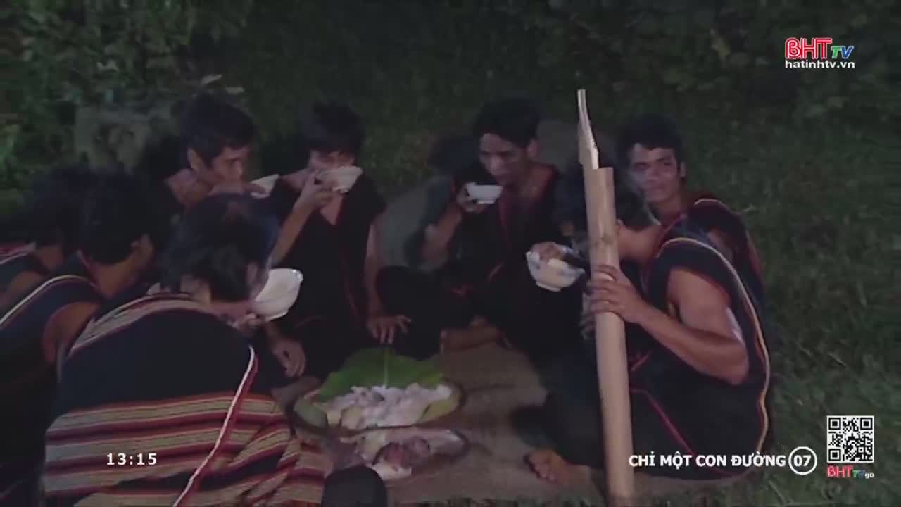 A group of men sit on the ground, eating from small bowls. One of them holds a long bamboo pole, while the others focus on their meal.
