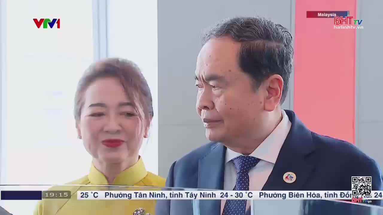 A woman in a yellow dress smiles, while a man in a suit looks off-camera. The scene appears to be from a broadcast on Ha Tinh TV, with the VTV1 logo visible.
