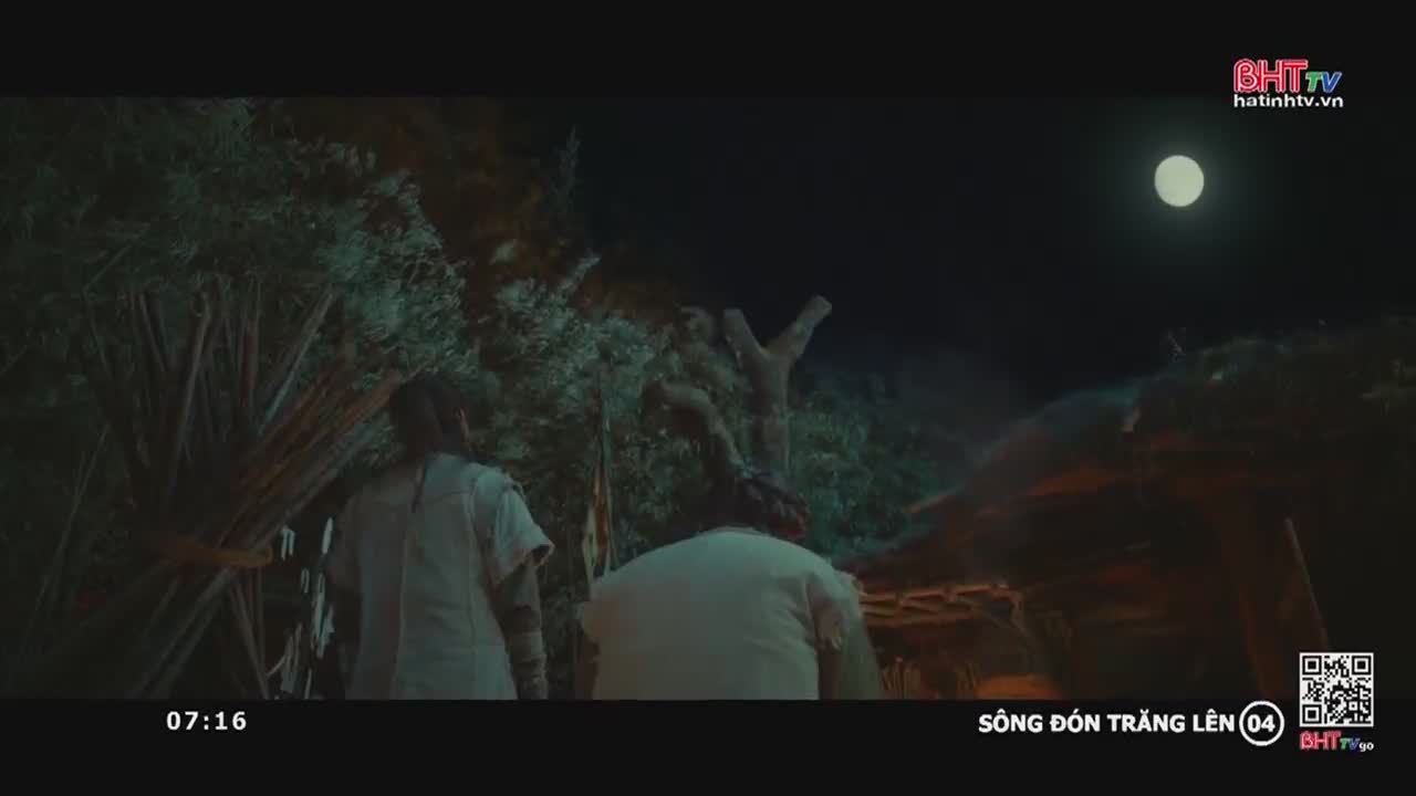 Two figures walk toward a small structure, the moon illuminating their path. One carries a bundle of branches, the scene broadcast on Ha Tinh TV in Vietnam.
