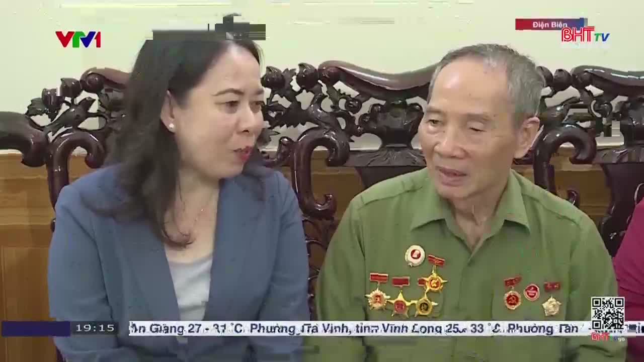 A woman in a blue blazer leans in, speaking intently to an older man in a green military-style shirt, covered in medals. The scene appears to be part of a broadcast on Vietnamese television, possibly Ha Tinh TV, with the time and weather displayed at the bottom of the screen.
