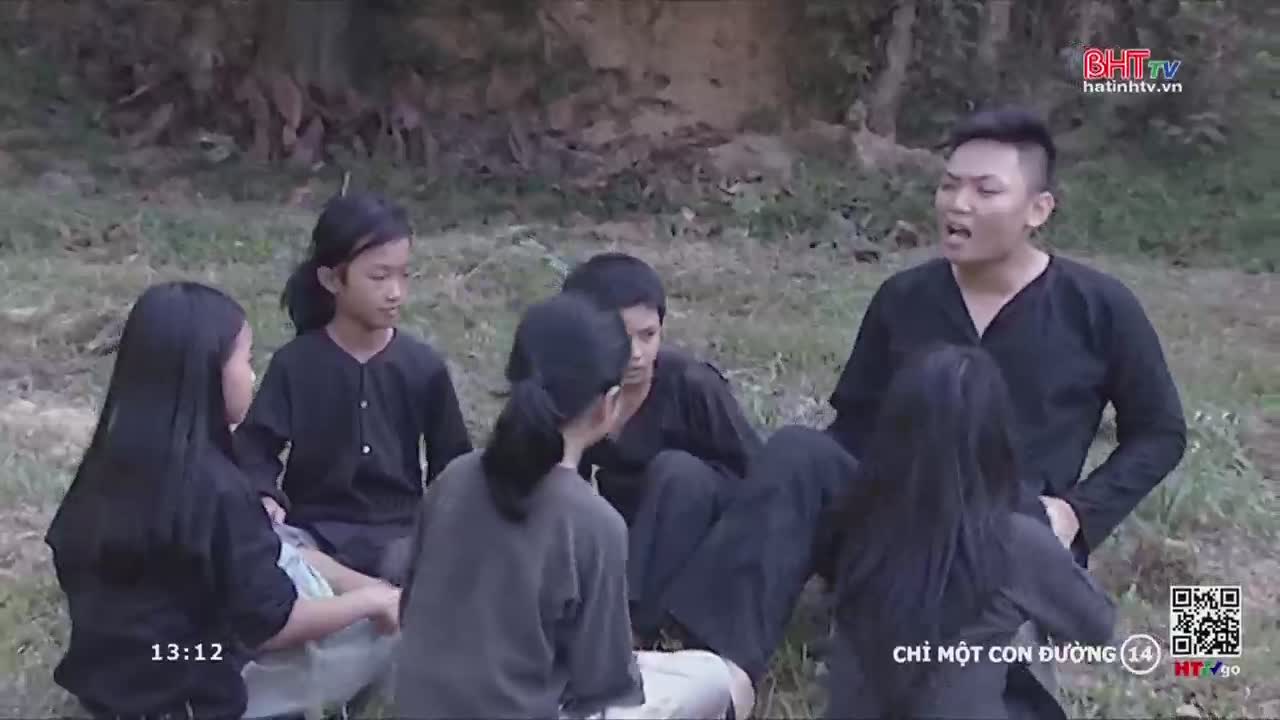 A group of young people in dark clothing are gathered on the grass, some are supporting the weight of another person. One man is speaking animatedly, likely directing the others.

