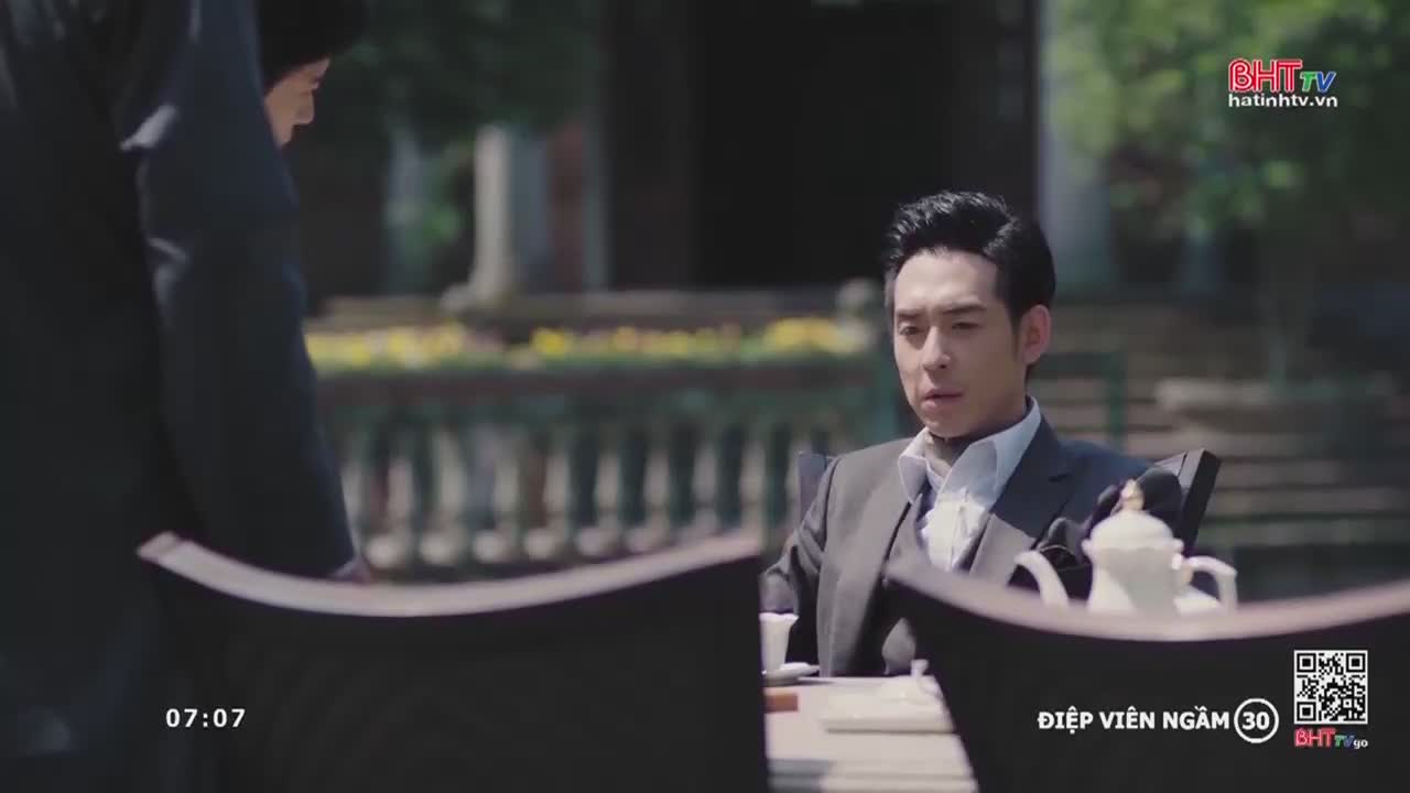 A man in a suit sits at a table, looking concerned as someone approaches him from the left.  This scene is from "Diep Vien Ngam" on Ha Tinh TV, airing in Vietnam.
