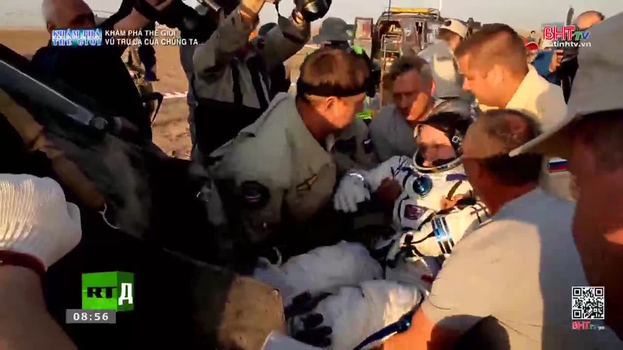 A cosmonaut in a white and blue suit is being helped out of a capsule by a group of people. The sun is low, casting long shadows across the dusty ground.