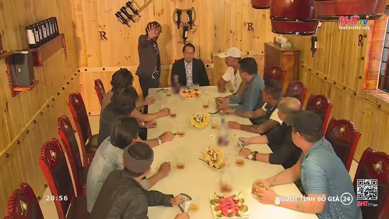 A man in a suit sits at the head of a long table, while others gather around him, some with drinks in hand. Another man stands to the side, gesturing with his hand.