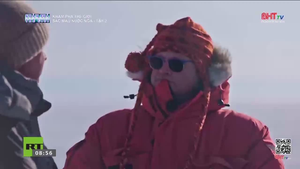 A man in a bright red parka and a patterned hat with ear flaps turns his head, his sunglasses reflecting the pale sky. Beside him, another person in a dark jacket looks off to the side. The BHT TV logo appears in the upper right corner.