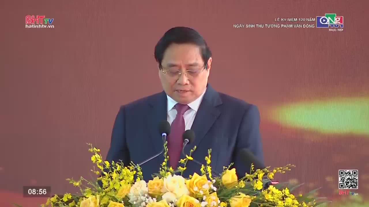A man in a dark suit stands at a podium, speaking into two microphones. He's surrounded by a vibrant arrangement of yellow and white flowers. The backdrop displays text, indicating a broadcast from Ha Tinh TV.