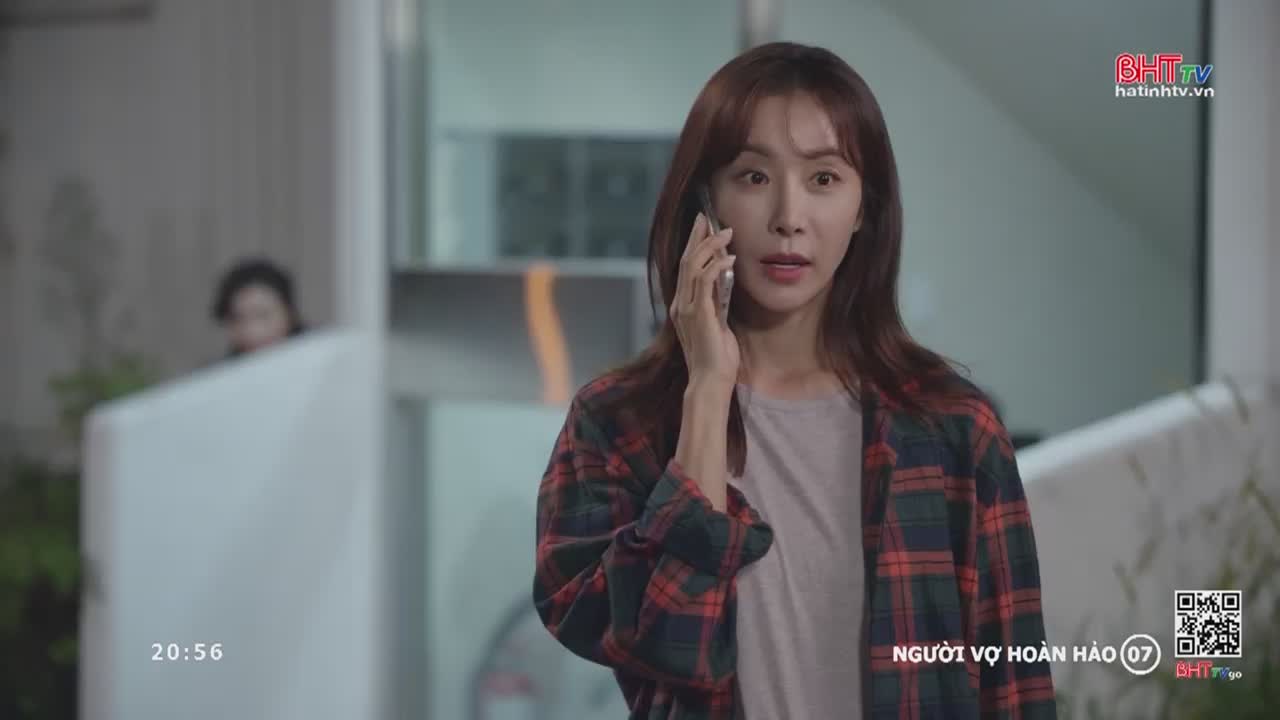 A woman with auburn hair holds a phone to her ear, her expression one of concern. Behind her, a blurred figure stands near a white partition.