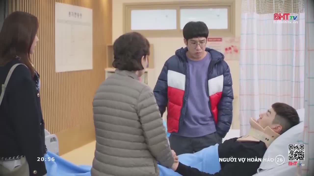 A young man lies in a hospital bed, his neck supported by a brace. A woman in a puffer jacket holds his hand, while another woman and a man in a colorful jacket stand nearby.