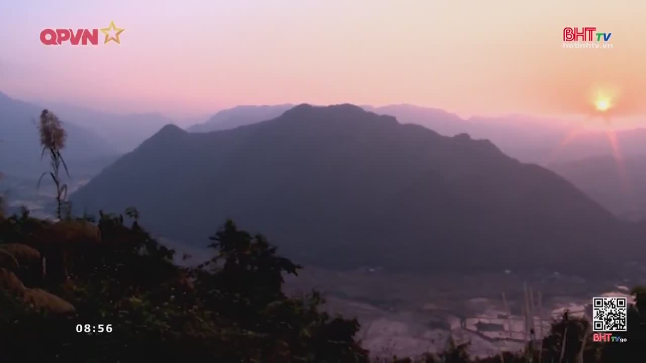 The sun crests over the hazy mountains of Vietnam, casting a soft pink glow across the valley. A lone stalk of tall grass stands silhouetted against the dawn sky.