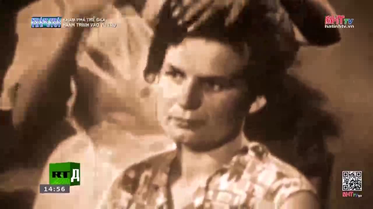 A woman with dark hair looks directly ahead, her expression unreadable. Her hands are clasped in her lap, and she wears a patterned blouse.