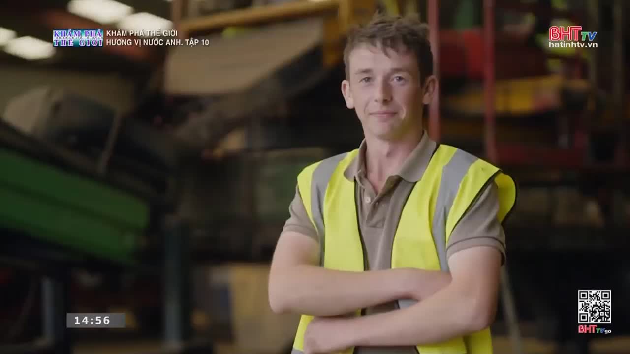 A young man stands with his arms crossed, a bright yellow safety vest over his shirt. Behind him, stacks of what look like car parts fill the background of this workshop.