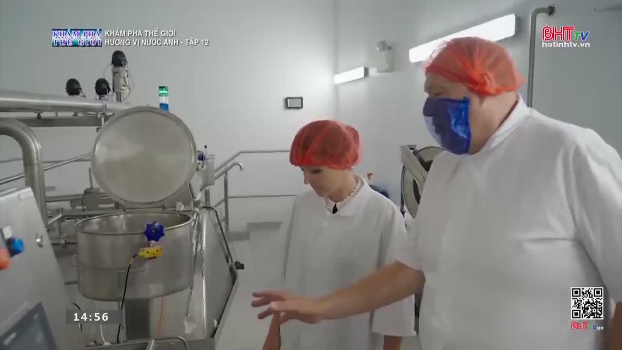 A man in a blue face shield and a woman in a red hairnet stand near a large, open stainless steel vat. The man gestures towards the equipment, explaining something about the process.