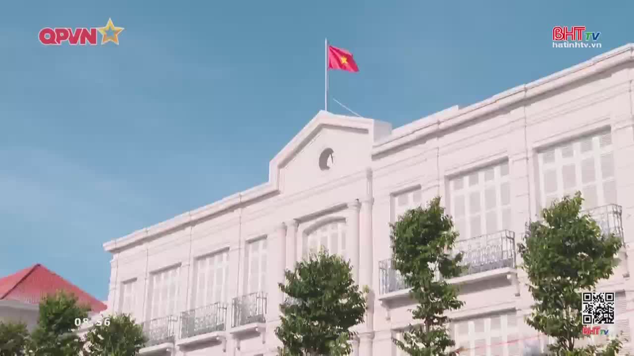The Vietnamese flag flies from a tall pole atop a grand, pale building. Trees with lush green leaves stand in front of the structure.
