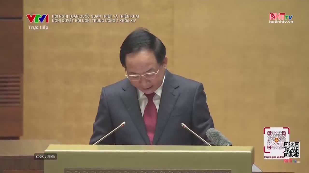 A man in a dark suit and red tie stands at a podium, speaking into a microphone. The screen displays "VTV Trực tiếp" and "BHTV hatinhtv.vn."