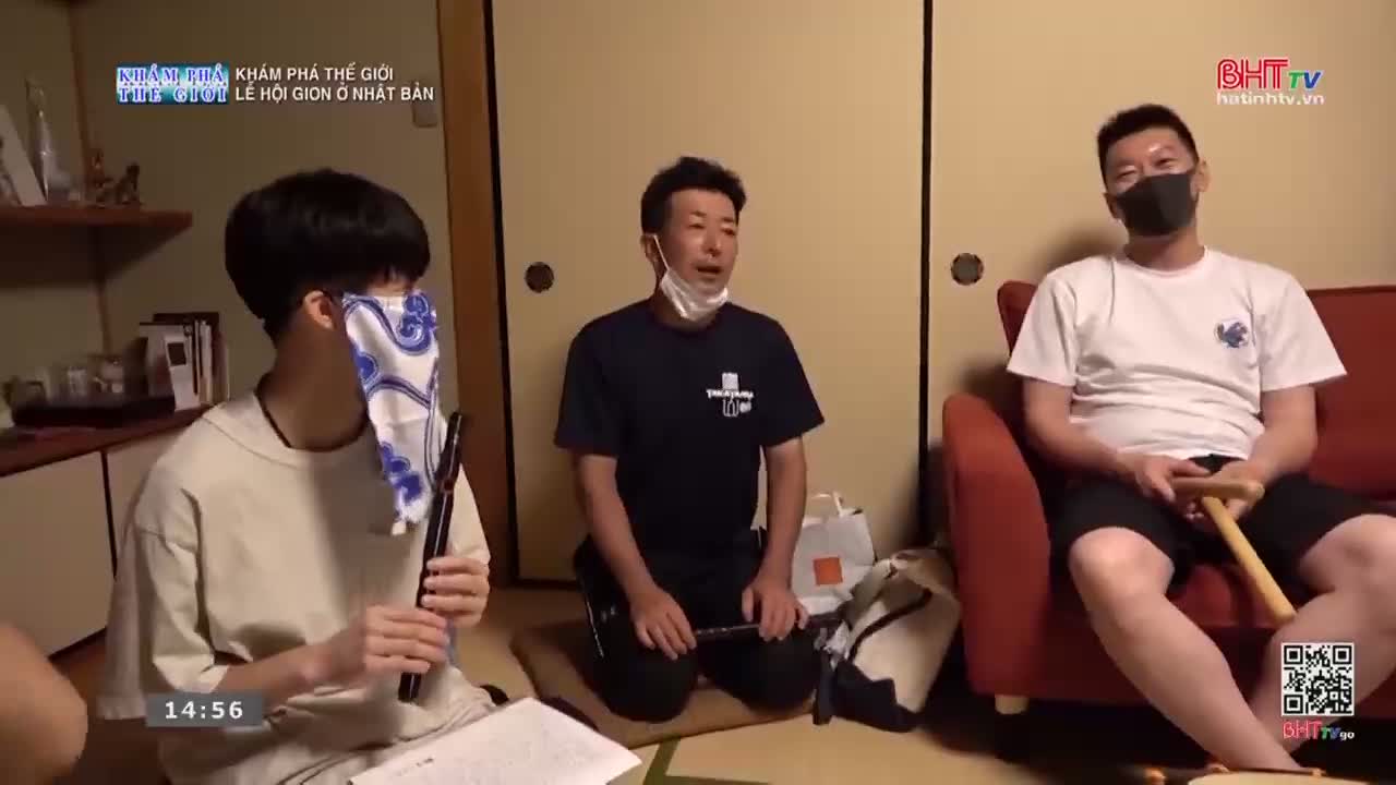 A young man, his eyes covered by a patterned cloth, holds a dark flute. Beside him, a man in a navy shirt kneels, his hands resting on a small stringed instrument. To their right, another man sits on a red couch, a black mask covering his face.