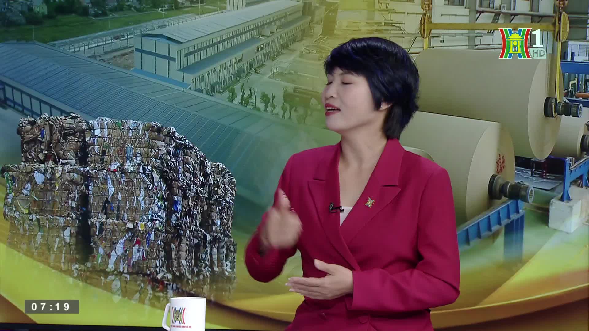 A woman in a red jacket gestures as she speaks, a stack of compressed paper and large rolls of paper are visible behind her. The HanoiTV1 logo is in the corner, and the time is displayed on the screen.
