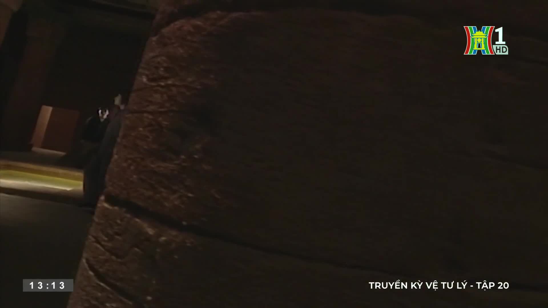 A figure in a dark suit stands in a dim doorway, partially obscured by a textured, brown wall. The HanoiTV1 logo and Vietnamese text indicate a scene from a television program.
