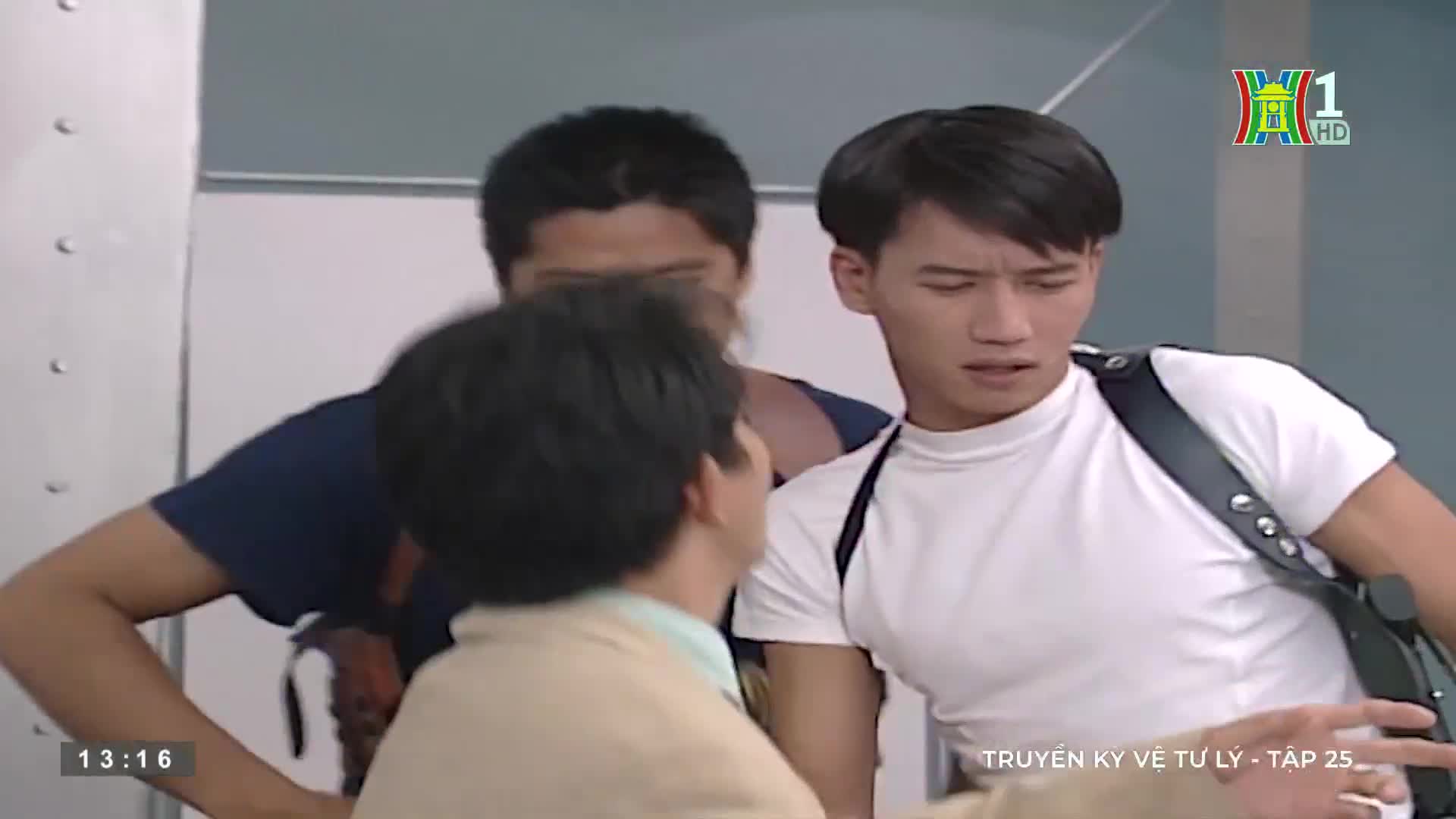 A man in a white t-shirt looks concerned, his eyes wide as he listens to someone off-screen. Two other men stand behind him, one with his arm resting on the shoulder of the man in the white shirt.
