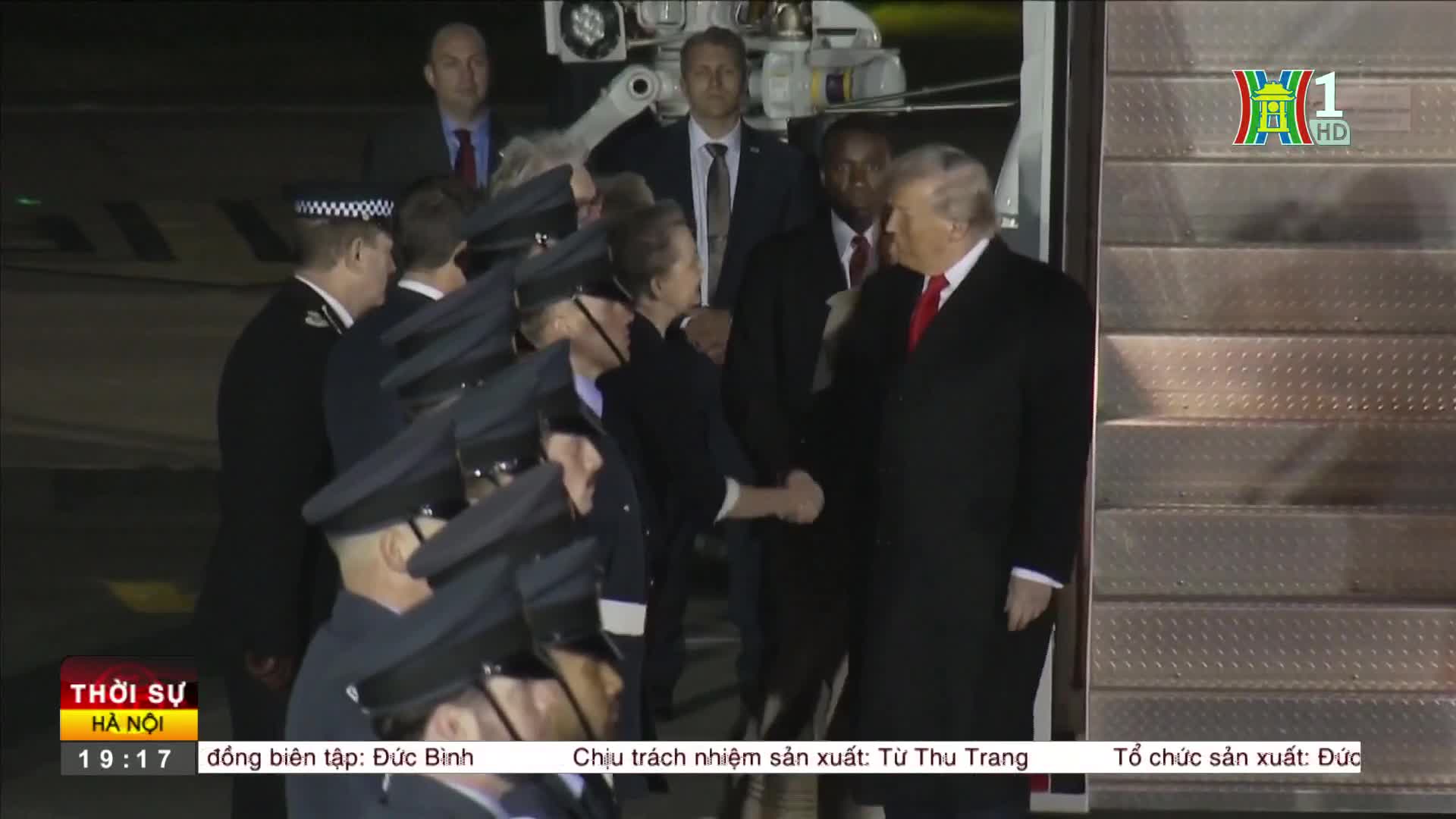 A man in a dark coat and red tie shakes hands with a person in a dark uniform, flanked by others in similar attire. The HanoiTV1 logo is visible as the scene unfolds.
