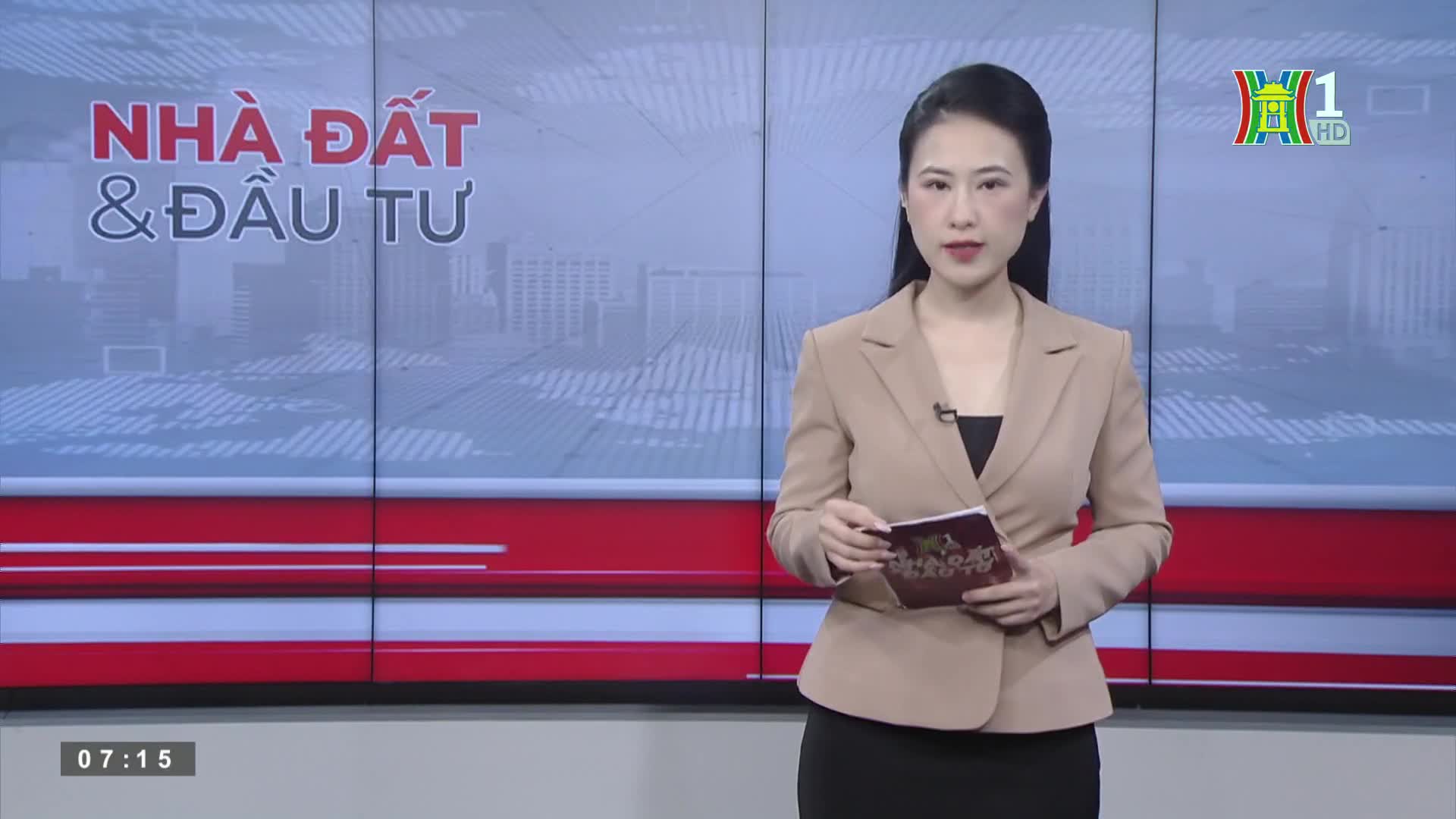 A woman in a beige blazer and black skirt stands center stage, holding a small card. Behind her, the HanoiTV1 logo is visible, along with the words "Nhà Đất & Đầu Tư."

