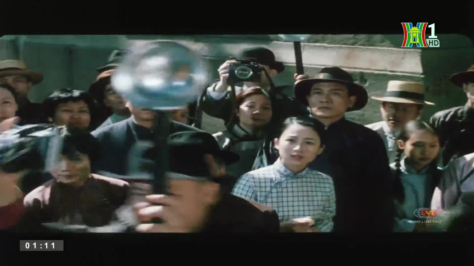 A crowd of people, many in hats, is gathered, some looking directly at the camera. A man holds a camera high, while another obscures the view with a large flashbulb.
