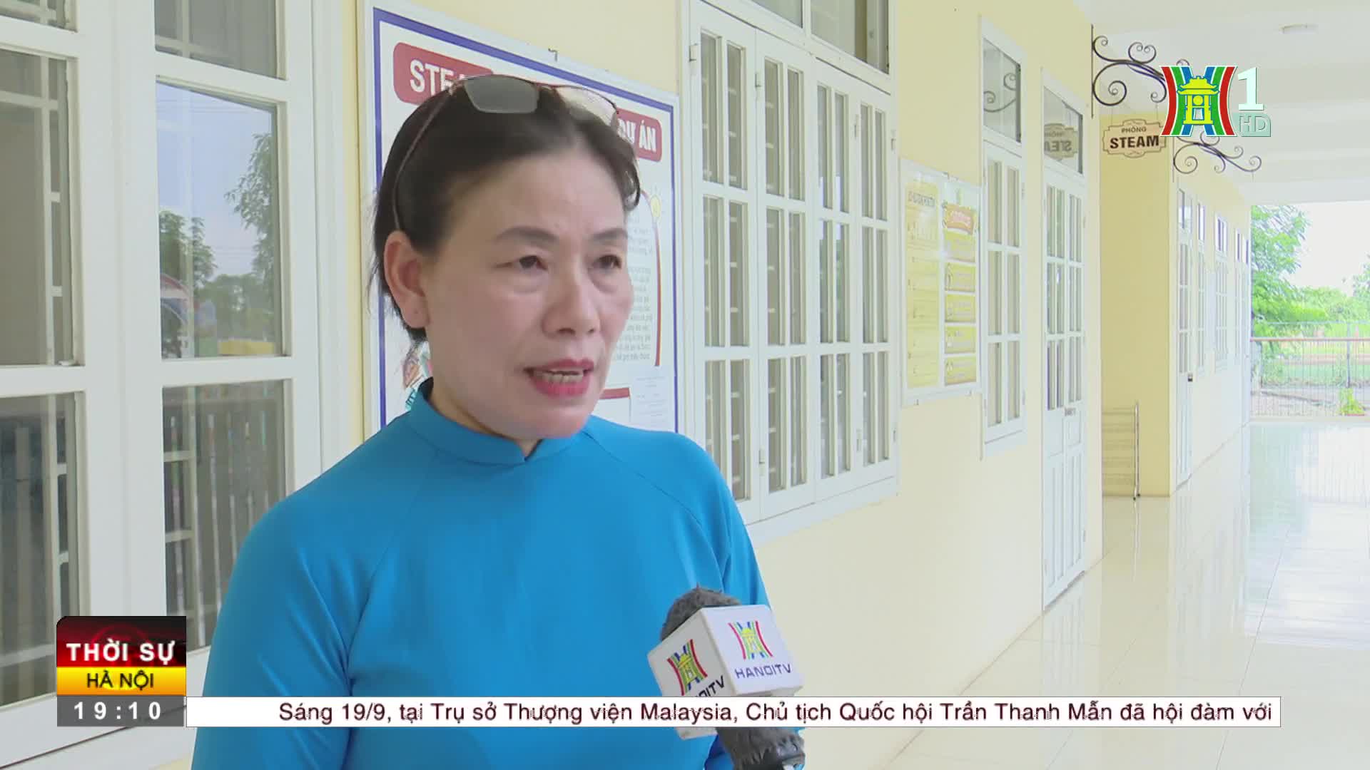 A woman in a blue shirt speaks into a HanoiTV1 microphone, her words broadcast on a screen. Behind her, sunlight streams through the windows of a building in Vietnam.
